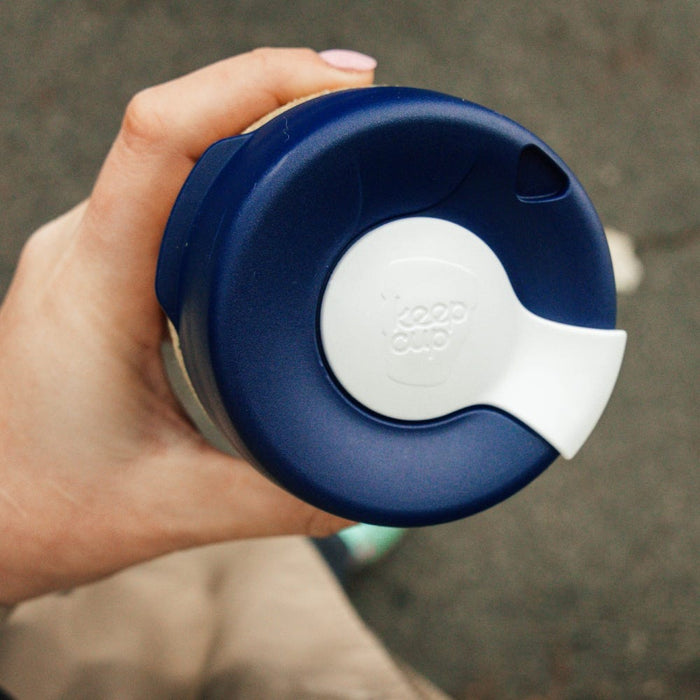Hand holding a KeepCup Brew Cork Ace in Rally colourway, viewed from above showing the navy blue lid with white flip-top closure, with a blurred dog visible in the background.