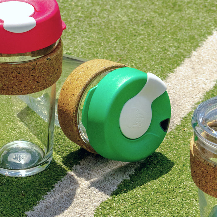 KeepCup Brew Cork Ace M 12oz in Slam green lies on its side on an artificial grass sports field, alongside other KeepCup variants with cork bands, showcasing the glass body, cork sleeve and colourful splash-proof lid.