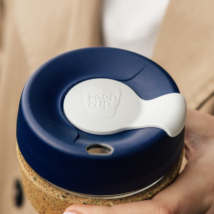 A hand holds the KeepCup Brew Cork Ace L 16oz, showing a close-up of the navy blue lid with white silicone sipper plug featuring the embossed KeepCup logo, with the natural cork band visible below.