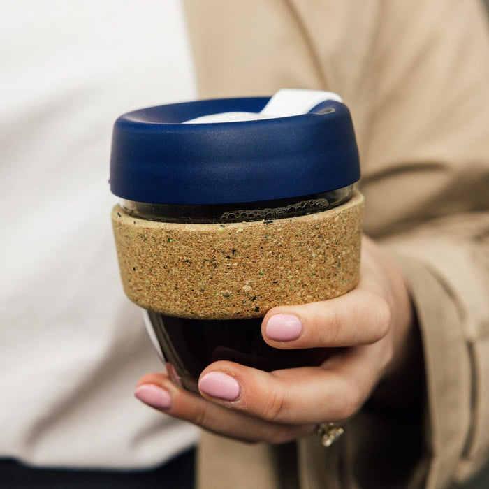 A hand with light pink nail polish holds a KeepCup Brew Cork Ace S 8oz in the Rally colourway, featuring a navy blue lid, natural cork band, and glass cup filled with dark coffee.