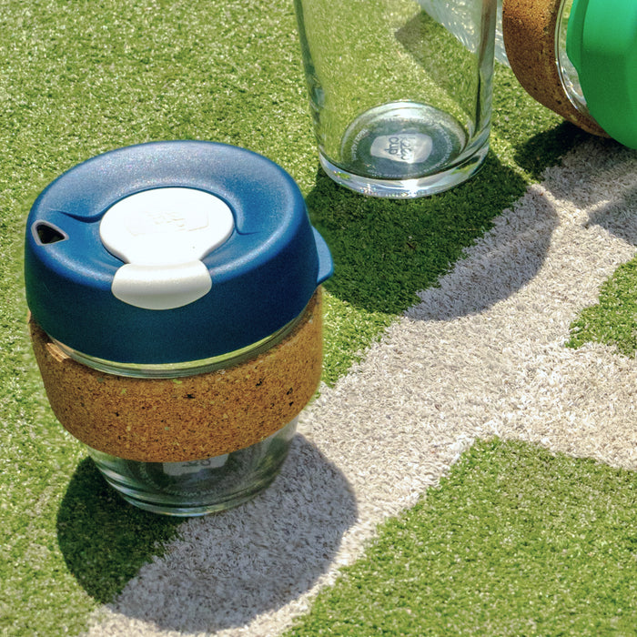 KeepCup Brew Cork Ace in size S 8oz, featuring a navy blue lid with white plug and natural cork band, sitting on artificial turf beside a white pitch marking with other reusable cups partially visible in the background.