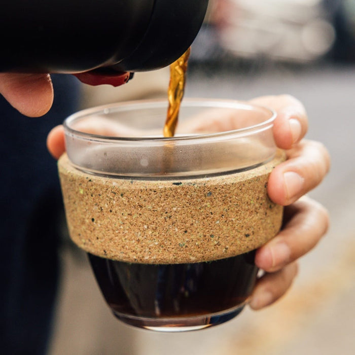 A hand holds a KeepCup Brew Cork Ace S 8oz in Slam colour while dark coffee is poured into it from a black vessel, showing the clear glass cup with natural cork band sleeve.