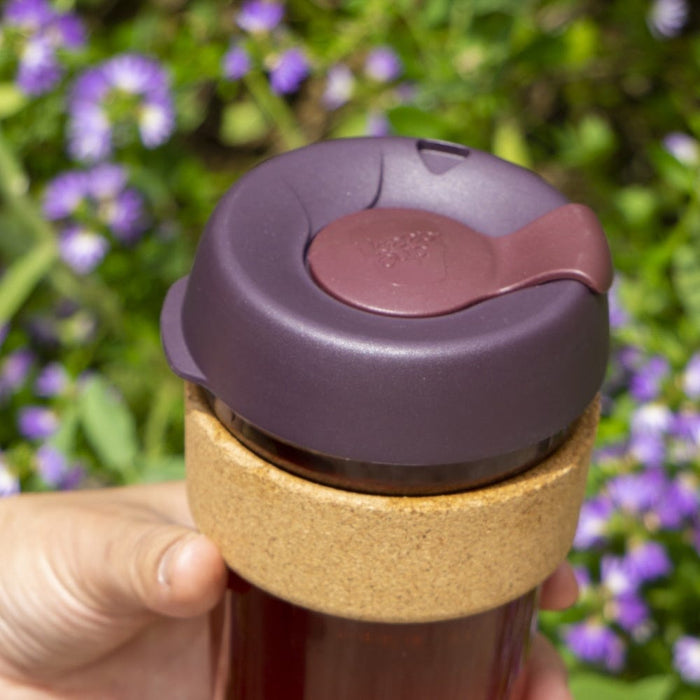 Hand holding a KeepCup Brew Cork M 12oz in Alder, featuring a deep plum-coloured lid with sipper closure and natural cork band, photographed outdoors against a backdrop of purple flowers and green foliage.
