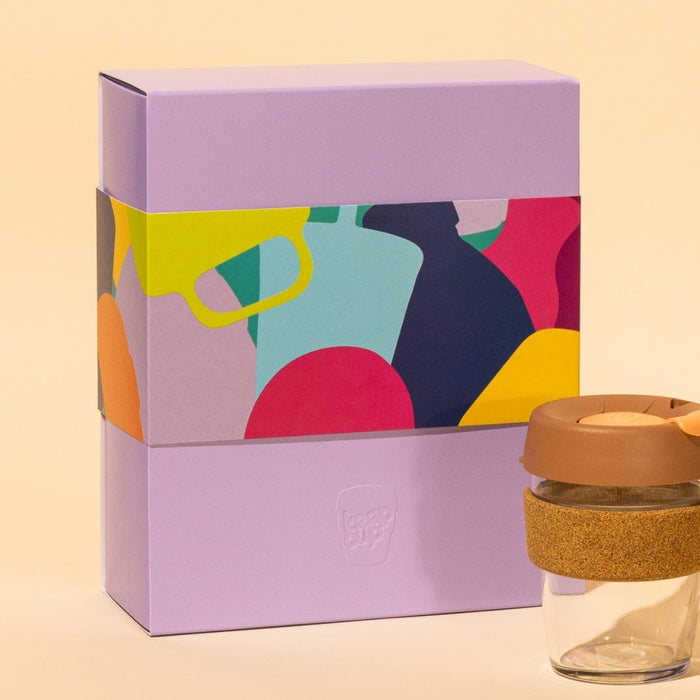 KeepCup Booked & Busy Alder reusable glass coffee cup with cork band and caramel-coloured lid, displayed next to its lavender gift box featuring a colourful abstract design of overlapping cup silhouettes.