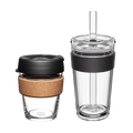 Two KeepCup reusable cups from the Booked & Busy Black set shown side by side against a white background: a smaller glass cup with cork band and black lid on the left, and a taller clear tumbler with black silicone band, clear lid and plastic straw on the right.