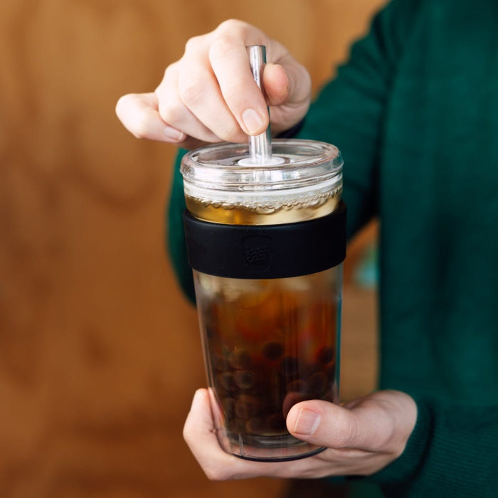 Hands holding a KeepCup Booked & Busy in Black, with a metal straw inserted through the clear lid. The reusable cup features a black silicone band and contains an iced coffee with visible ice cubes.
