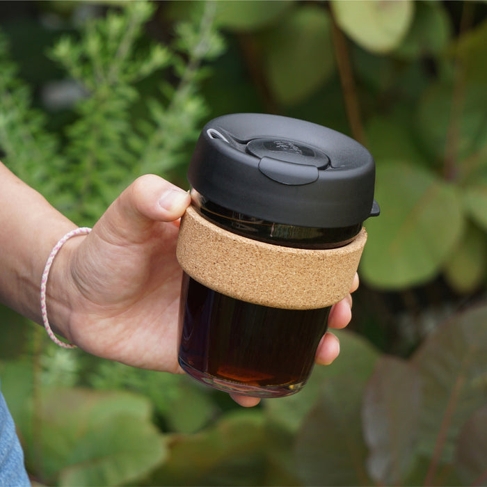 A hand holds the Booked & Busy KeepCup in Black, featuring a glass body filled with dark coffee, natural cork band, and black lid with flip-top closure, photographed outdoors against a backdrop of green foliage.