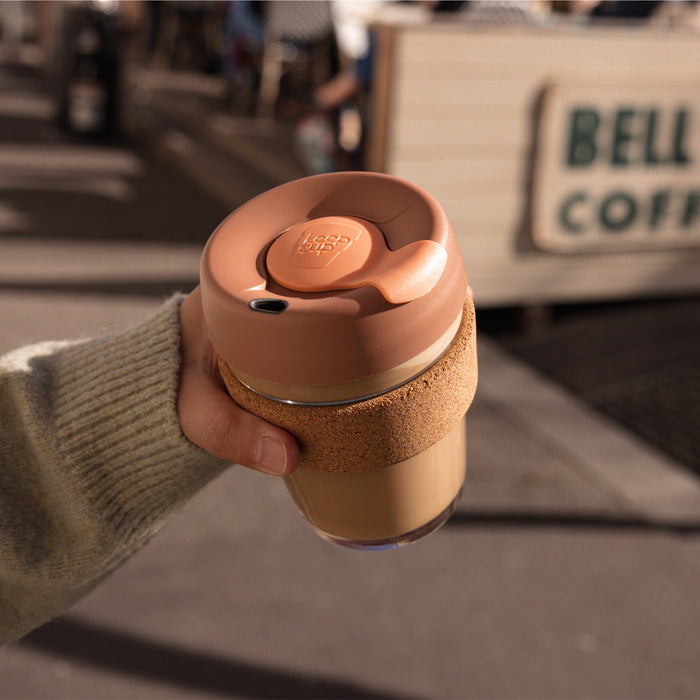 A hand in an olive green knit jumper holds a KeepCup Booked & Busy in Frappe, featuring a glass cup with cork band and peach-coloured lid, with a coffee cart visible in the blurred background.