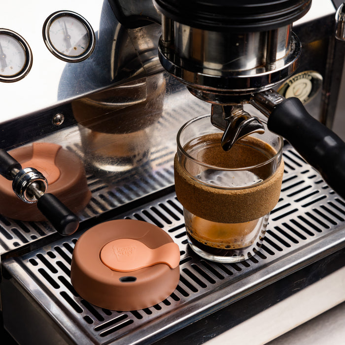 KeepCup Brew Cork M 12oz in Frappe colour positioned under an espresso machine group head with fresh coffee being extracted, the terracotta-coloured silicone lid resting beside it on the stainless steel drip tray.