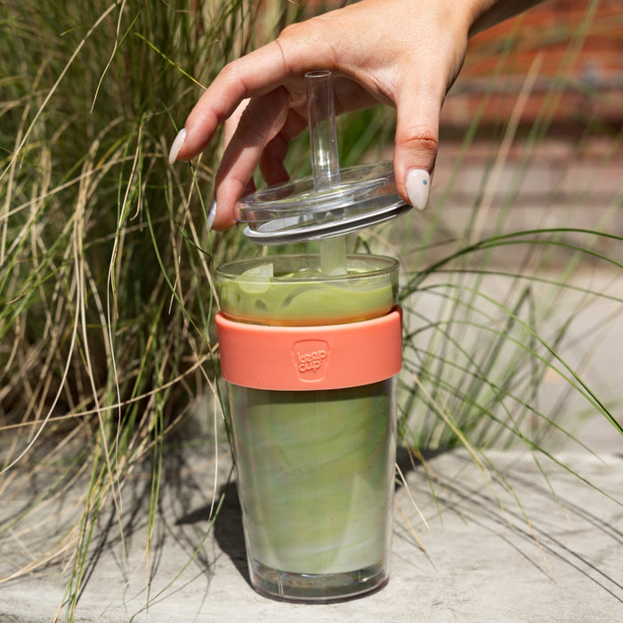 A hand reaches to lift the clear lid with straw from a KeepCup Cold Cup Longplay Boba L 16oz in Clay, filled with iced green matcha drink, set against ornamental grasses outdoors.