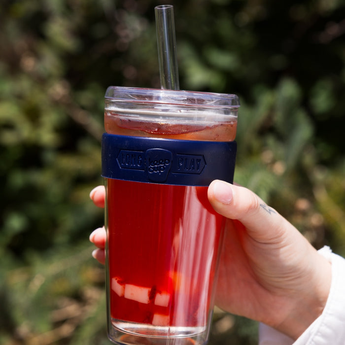 Hand holding a KeepCup Cold Cup Longplay Boba L 16oz in Navy, filled with iced red beverage and featuring a clear straw, navy silicone band, and transparent lid, photographed outdoors against a blurred green foliage background.