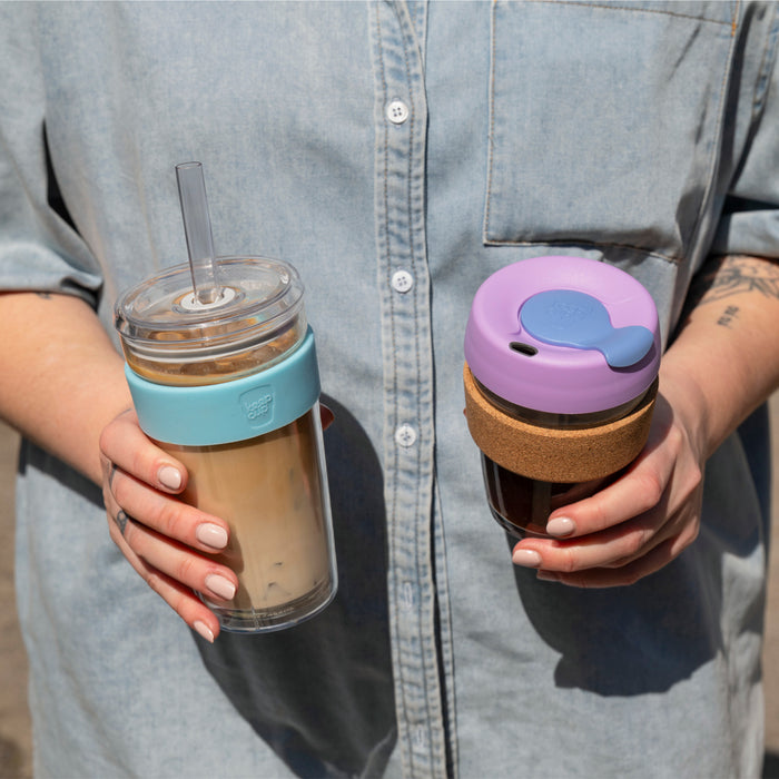 A person in a light denim shirt holds two KeepCup reusable cups — the left hand holds a clear tumbler with iced coffee and a teal band featuring a straw, while the right hand holds the Booked & Busy Moonlight KeepCup with a lavender lid, blue plug, and cork band filled with dark coffee.