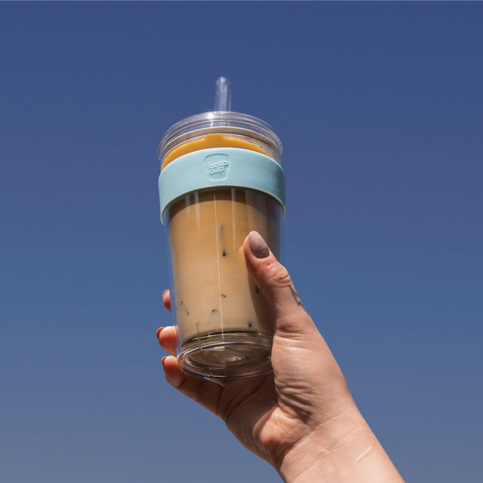 Hand with orange nail polish and a small tattoo holds up a KeepCup Booked & Busy in Moonlight colour against a clear blue sky, the reusable cup filled with iced coffee and featuring a light blue silicone band, clear straw, and double-walled design.