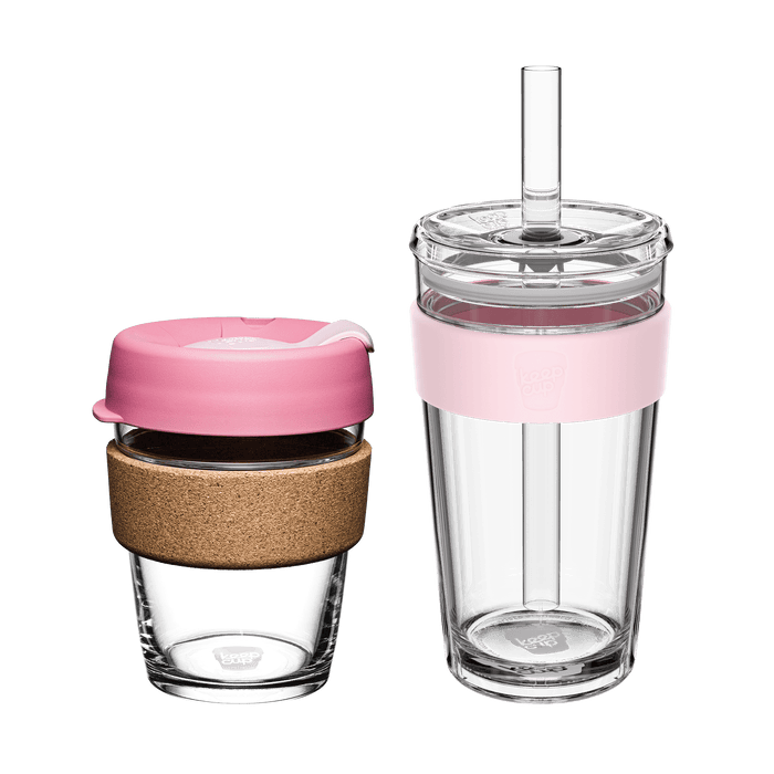 Two KeepCup reusable cups in the Booked & Busy Pashmak Pink colourway displayed side by side on a white background: a glass coffee cup with cork band and pink plastic lid, and a taller clear tumbler with pink silicone band, clear lid and plastic straw.