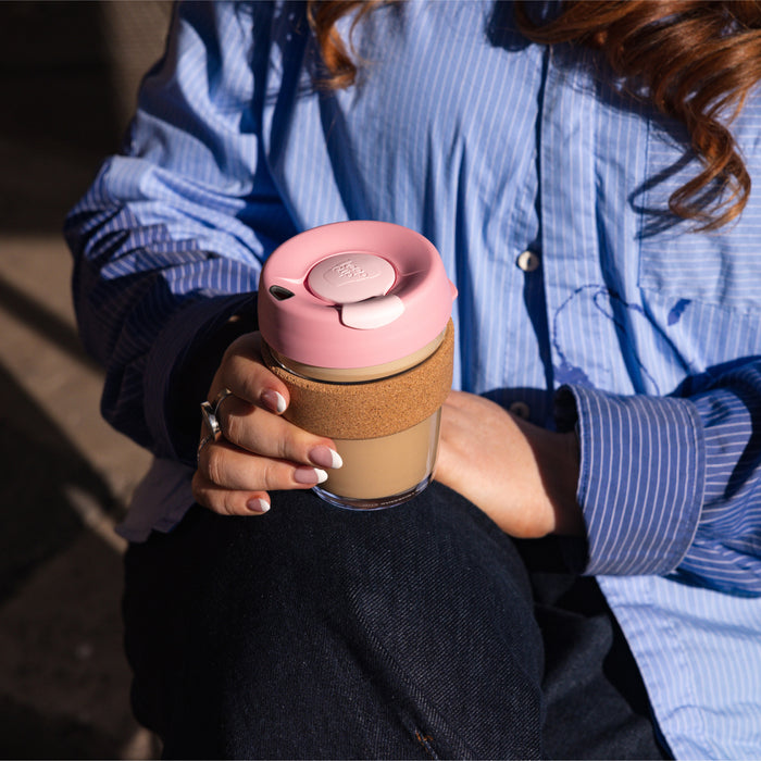 A woman in a blue striped shirt sits outdoors holding a KeepCup Booked & Busy in Pashmak Pink, featuring a glass cup with cork band and pastel pink lid, filled with a creamy coffee drink.