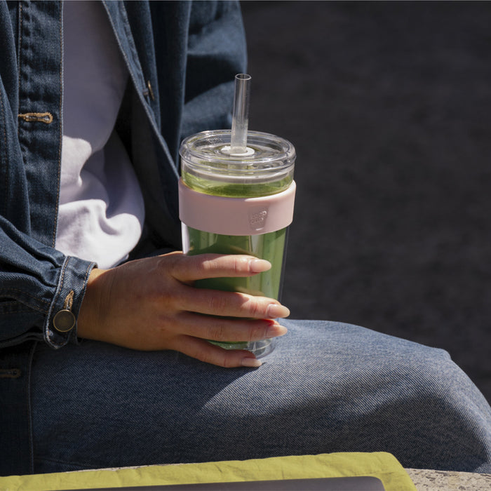 A person in a denim jacket sits outdoors holding a KeepCup Booked & Busy in Pashmak Pink filled with a green smoothie, the clear reusable cup featuring a soft pink silicone band and a straw lid.