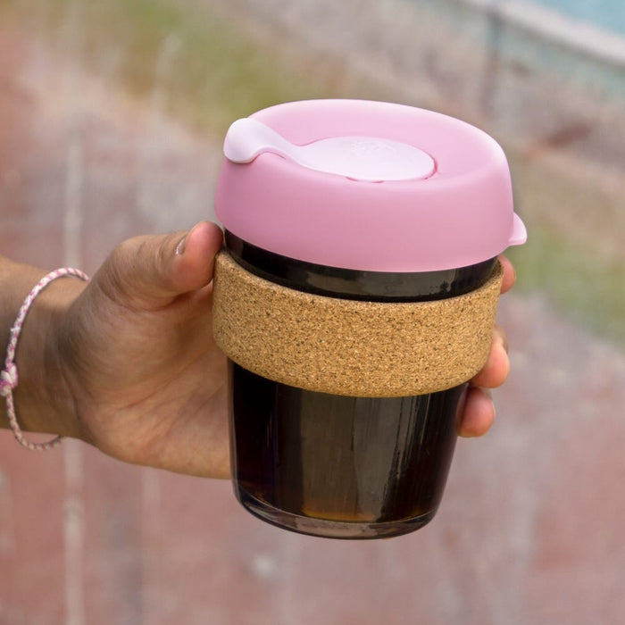 A hand holding a KeepCup Booked & Busy reusable coffee cup in Pashmak Pink, featuring a tinted glass body with dark liquid inside, natural cork band sleeve, and pastel pink plastic lid with a flip-top closure, photographed outdoors against a softly blurred background.