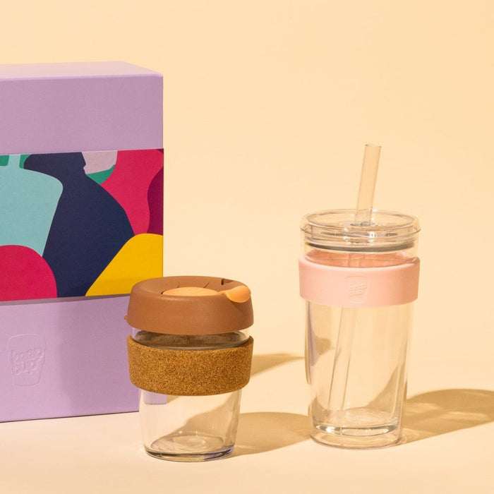 KeepCup Booked & Busy gift set featuring a glass reusable coffee cup with cork band and brown lid alongside a clear tumbler with pink silicone band and straw, displayed next to a colourful purple gift box on a warm cream background.