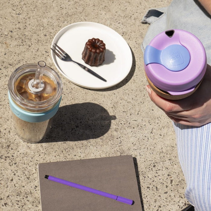A hand holds a KeepCup Booked & Busy in Moonlight, featuring a lavender lid and cork band, viewed from above on a sunlit concrete surface alongside an iced coffee in a teal KeepCup, a canelé on a white plate, and a grey notebook with purple pen.