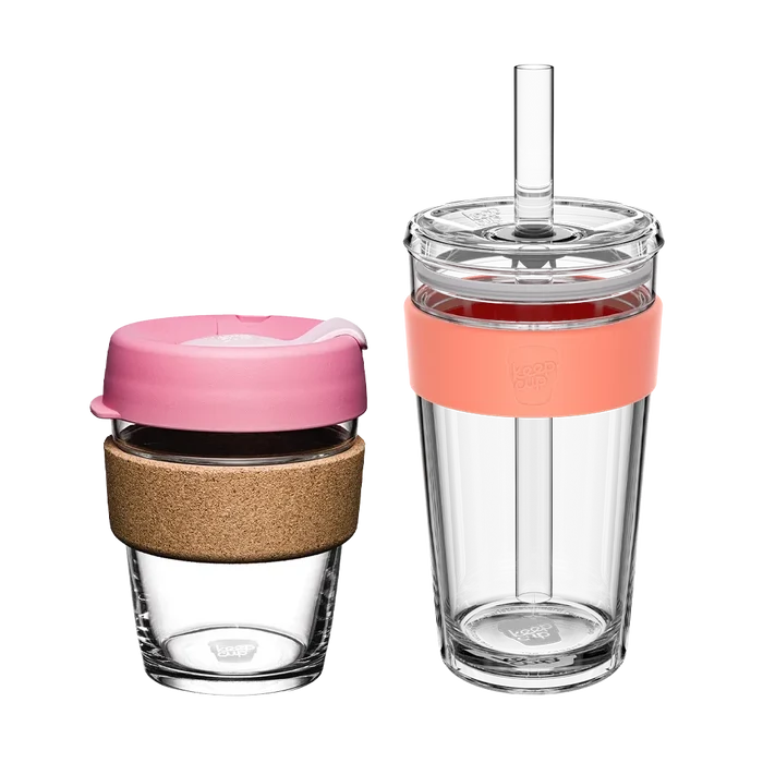 Two KeepCup reusable cups from the Booked & Busy Saskatoon set displayed side by side: a smaller glass cup with cork band and pink lid on the left, and a taller clear tumbler with peach silicone band and straw on the right, both shown against a white background.