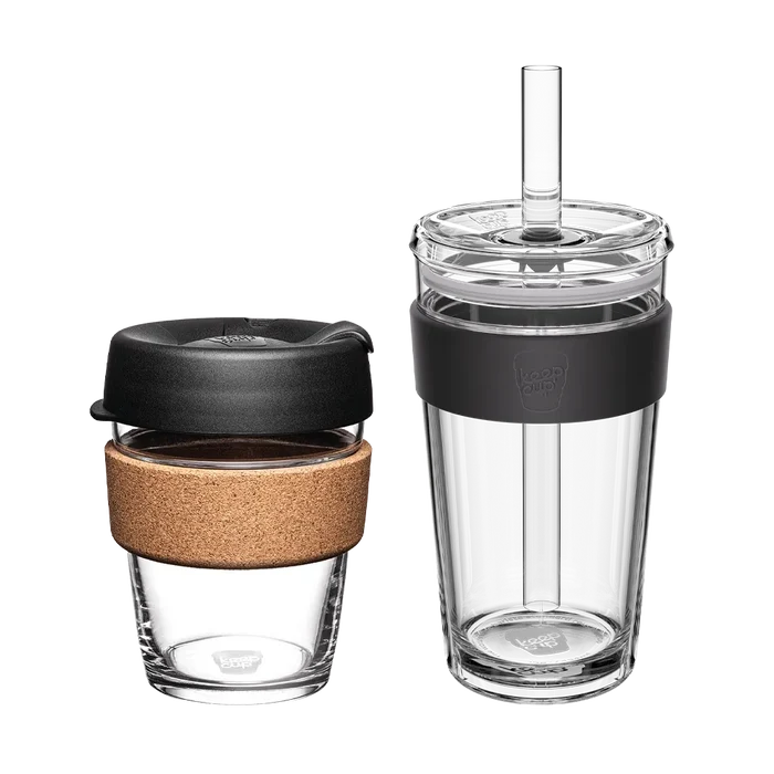 Two KeepCup reusable cups from the Booked & Busy Black set shown side by side against a white background: a smaller glass cup with cork band and black lid on the left, and a taller clear tumbler with black silicone band, clear lid and plastic straw on the right.