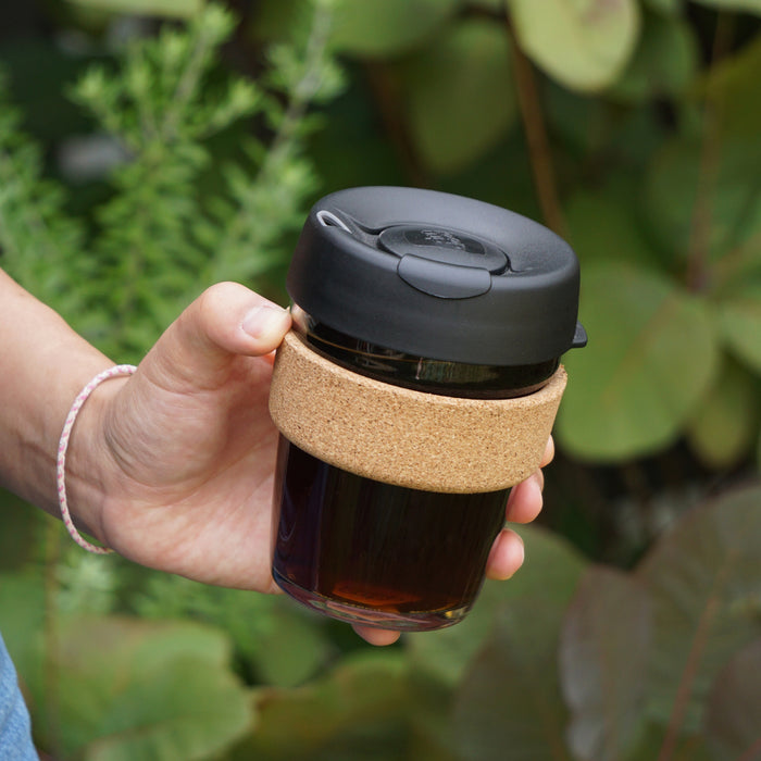 Hand holding a Brew Cork M 12oz reusable coffee cup in Black, featuring a glass body with dark liquid inside, natural cork band sleeve, and black plastic lid, photographed outdoors against a leafy green background.