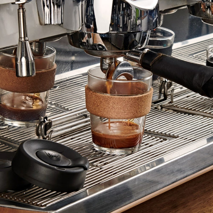 KeepCup Brew Cork M 12oz in Black sitting on an espresso machine drip tray, with fresh espresso being extracted into the glass cup, showcasing the cork band and black lid resting nearby.