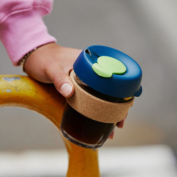 A hand wearing a pink sleeve holds a KeepCup Brew Cork M 12oz in Deep, featuring a navy blue lid with green plug, natural cork band, and glass cup filled with dark coffee, resting against a yellow metal railing.