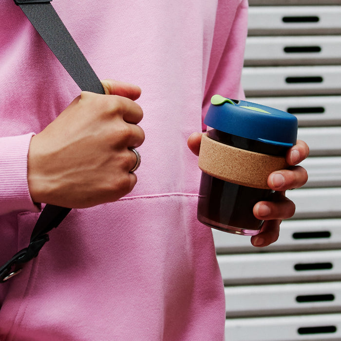 A person in a pink hoodie holds the KeepCup Brew Cork M 12oz in Deep, showcasing its dark glass body, natural cork band, and navy blue lid with green plug, while gripping a bag strap with their other hand.