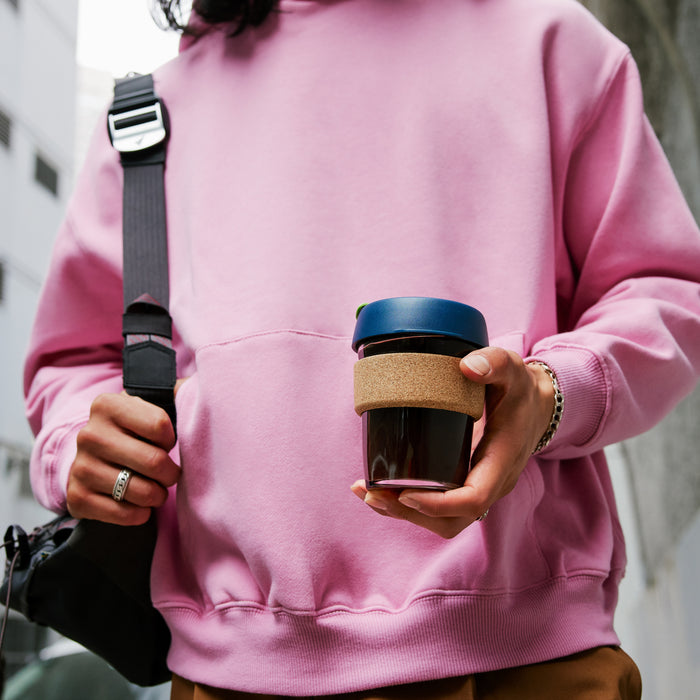 A person in a pink sweatshirt holds the KeepCup Brew Cork M 12oz in Deep, showcasing the dark glass cup with cork band and navy lid, while carrying a camera strap over their shoulder in an urban setting.