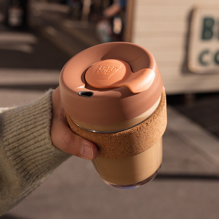 A hand wearing a khaki knit sweater holds a KeepCup Brew Cork M 12oz in Frappe, showing the peachy-pink lid with sipper plug and natural cork band wrapped around the glass cup filled with coffee, photographed outdoors in warm afternoon light.
