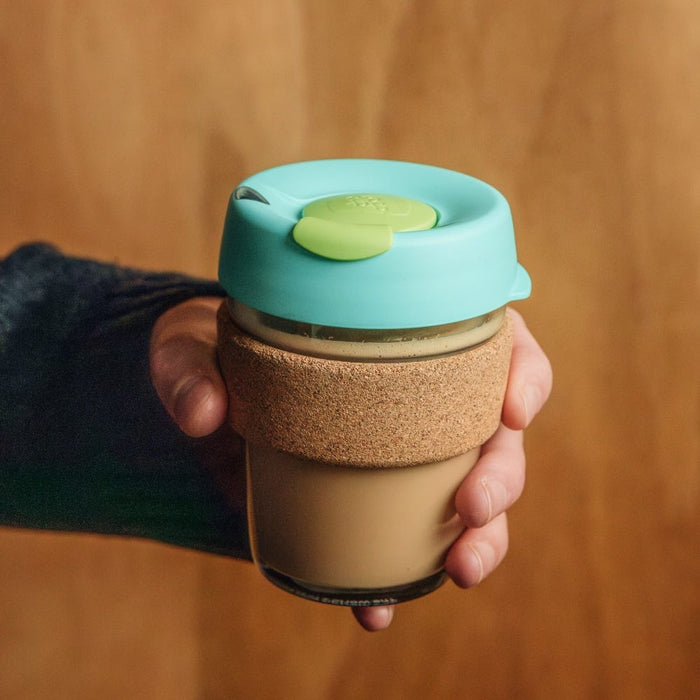 A hand holding a KeepCup Brew Cork M 12oz in Matcha, featuring a turquoise silicone lid with lime green plug, cork band sleeve, and tempered glass cup filled with a creamy latte, against a warm wooden background.