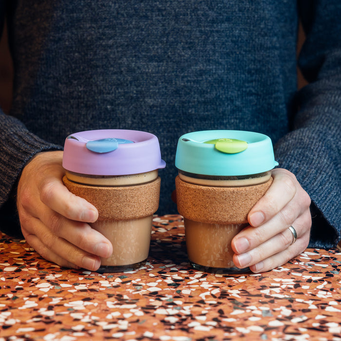 Two hands holding KeepCup Brew Cork reusable coffee cups side by side on a terrazzo table, featuring the M 12oz Matcha with its distinctive turquoise silicone lid and cork band, alongside a lavender-lidded variant, both filled with coffee.