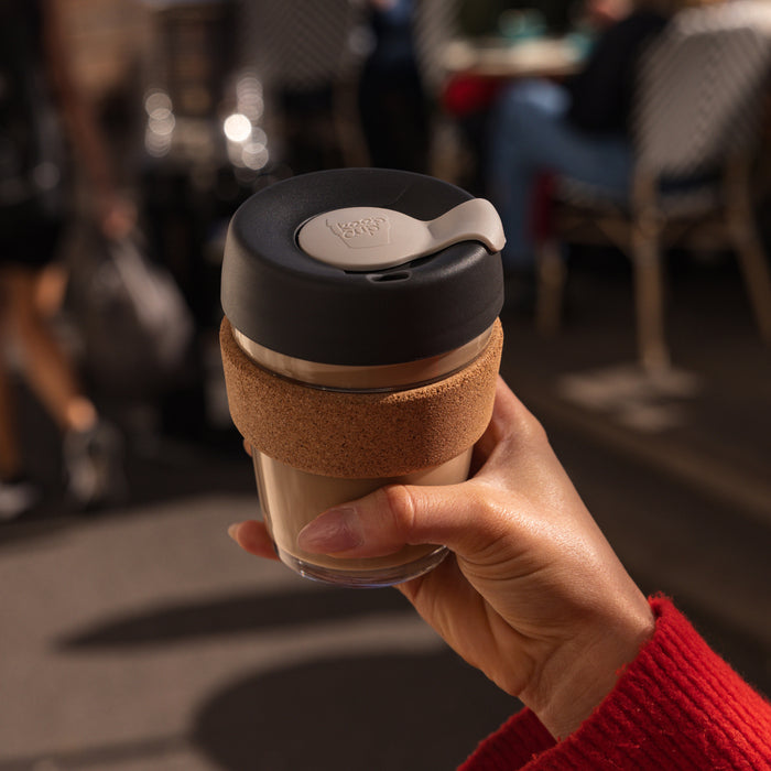 Hand holding a KeepCup Brew Cork M 12oz in Press colour, featuring a dark grey lid with light grey plug, natural cork band, and glass cup filled with a latte, with a blurred café setting in the background.