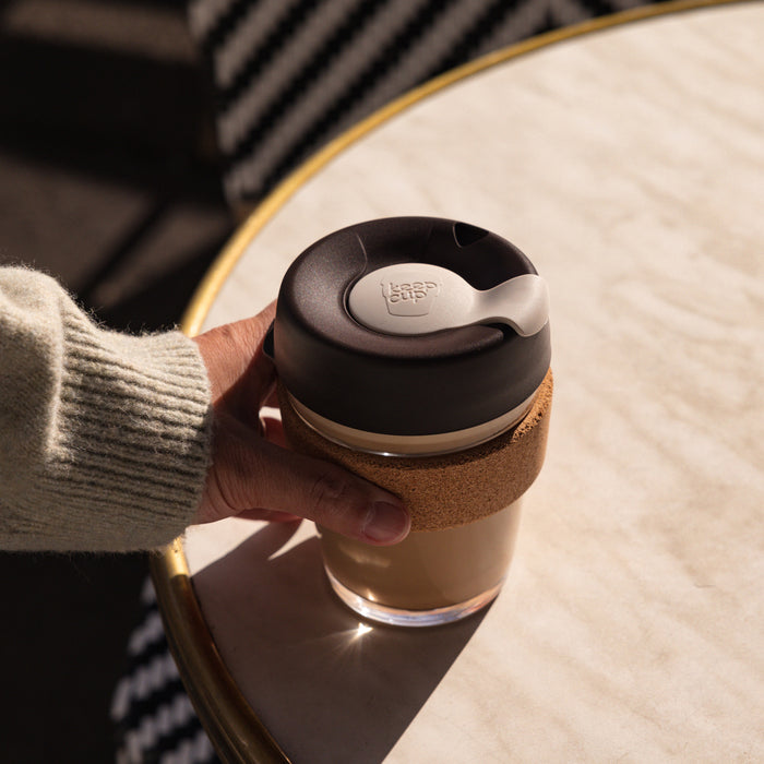 A hand in a cosy grey knit sweater holds a KeepCup Brew Cork M 12oz in Press, featuring a glass cup with cork band and dark lid, positioned over a marble café table in warm natural light.