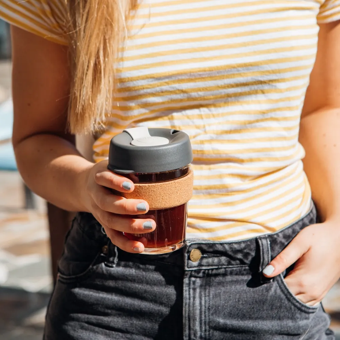 A woman in a yellow and white striped t-shirt holds a Brew Cork M 12oz KeepCup in Press colour, featuring a glass cup with cork band and dark grey lid, with black coffee visible inside.