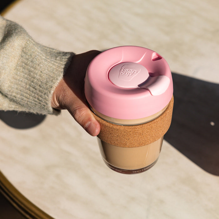 Hand holding a KeepCup Brew Cork M 12oz in Saskatoon, featuring a pink lid, natural cork band and glass cup filled with a creamy coffee, photographed from above against a light marble surface in natural sunlight.