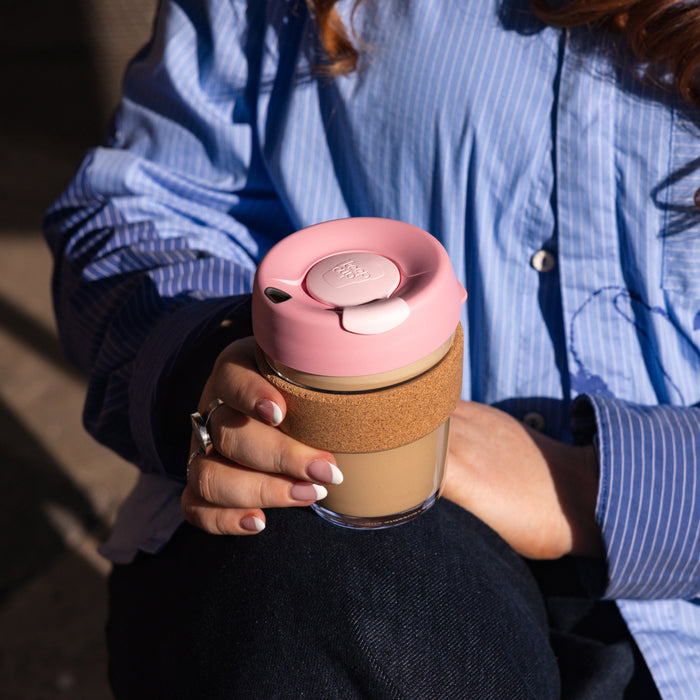A woman in a blue striped shirt holds a KeepCup Brew Cork M 12oz in Saskatoon, featuring a pink lid, natural cork band, and glass cup filled with a latte, photographed from above while she sits outdoors.