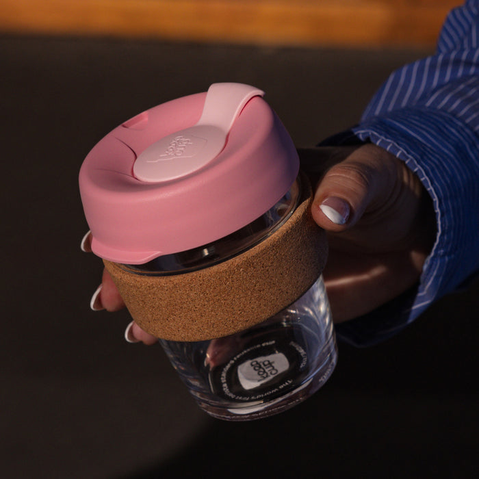 Hand holding a KeepCup Brew Cork M 12oz in Saskatoon, featuring a pink lid, tempered glass cup, and natural cork band, photographed from above against a dark background.