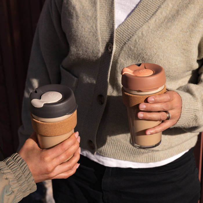 Two people holding KeepCup Brew Cork reusable coffee cups with cork bands, one in Frappe colour with a dusty pink lid, both filled with milky coffee, in a casual outdoor setting.