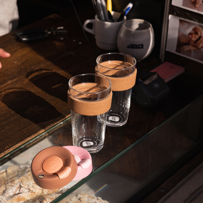 Two KeepCup Brew Cork L 16oz cups in Frappe sit on a glass café counter with their cork bands visible, alongside the pink and terracotta coloured lids removed and placed in front.