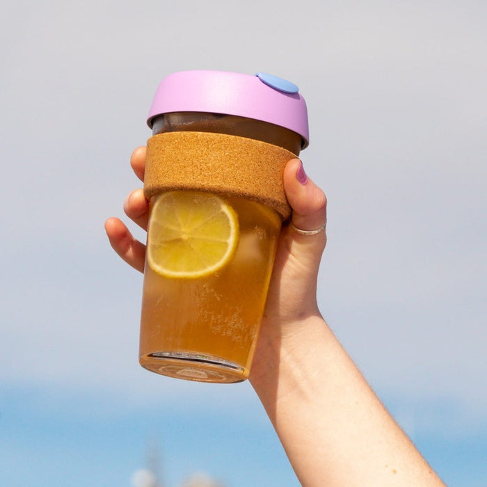Hand holding a KeepCup Brew Cork L 16oz in Moonlight, featuring a lavender silicone lid with blue plug, cork band, and glass cup filled with iced tea and a lemon slice, against a soft blue sky background.
