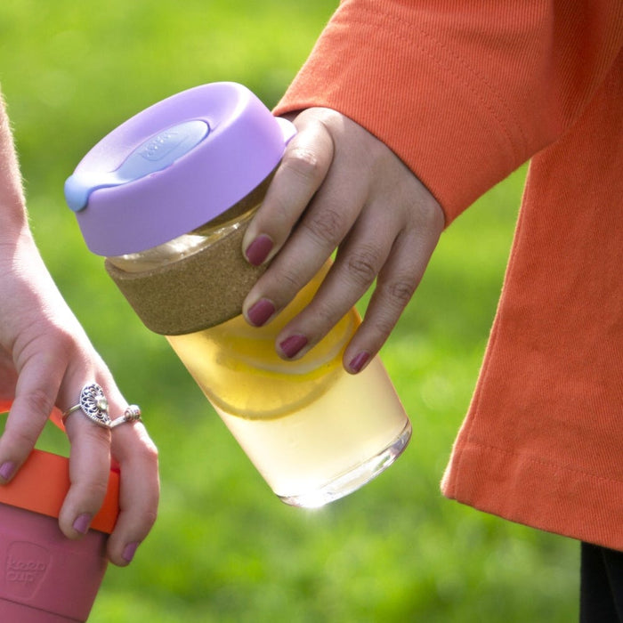 A hand with burgundy nail polish holds a KeepCup Brew Cork L 16oz in Moonlight, featuring a lavender lid, cork band, and glass body filled with a yellow beverage, against a grassy outdoor background with another person holding a pink KeepCup nearby.