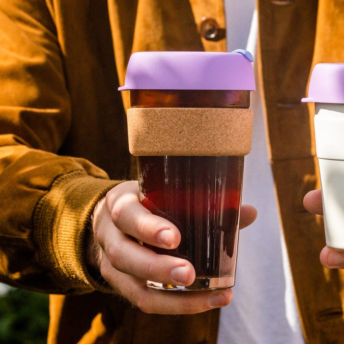 A hand holds the KeepCup Brew Cork L 16oz in Moonlight, showcasing the tinted glass cup filled with dark coffee, natural cork band, and lavender silicone lid, with a second person holding a white KeepCup visible in the background.