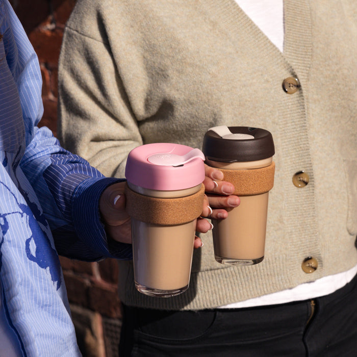 Two people holding KeepCup Brew Cork reusable coffee cups filled with milk coffee, featuring cork band sleeves — one with a pink Saskatoon lid and one with a dark lid — in a casual outdoor setting.