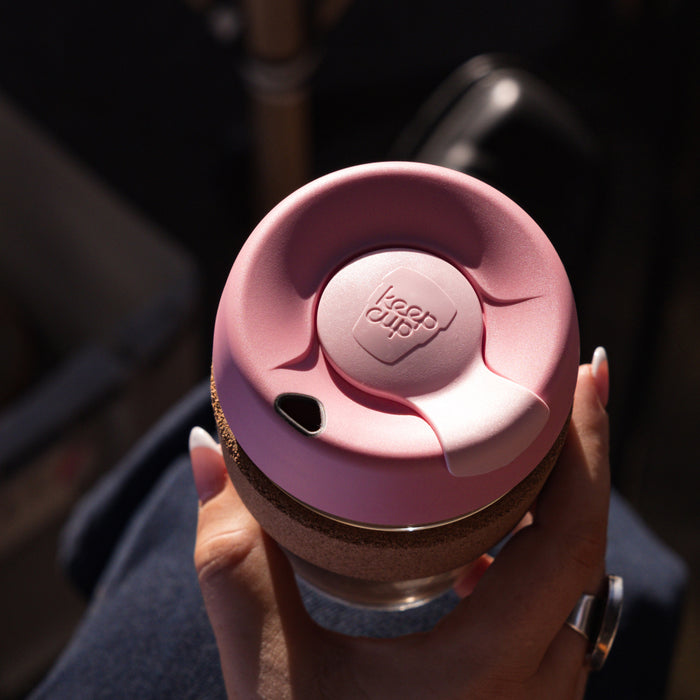 A hand with white nail polish holds a KeepCup Brew Cork L 16oz in Saskatoon, viewed from above showing the dusty pink lid with the KeepCup logo, cork band, and glass cup beneath.