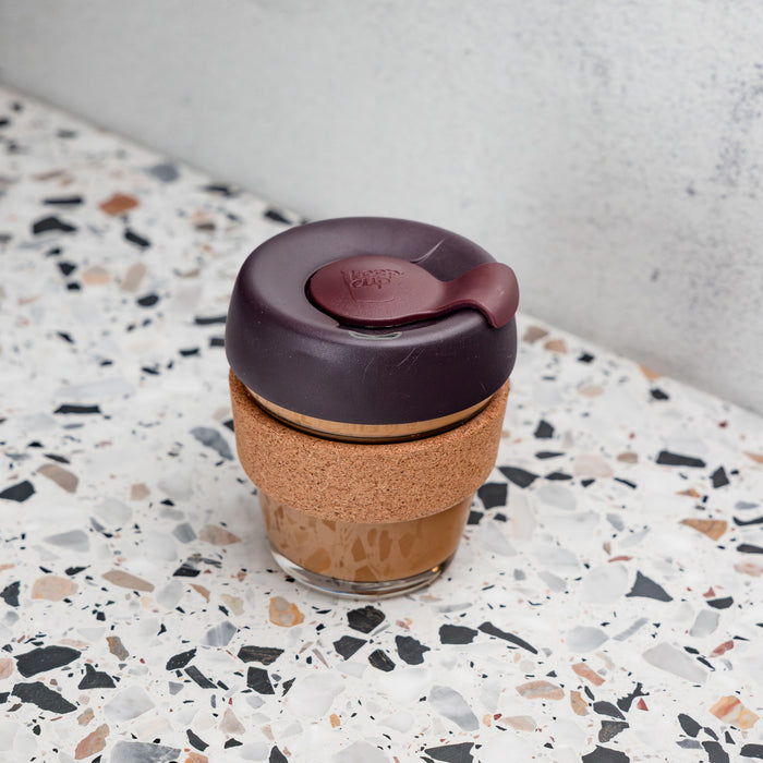 KeepCup Brew Cork XS 6oz in Alder, featuring a glass cup with cork band and deep plum-coloured lid, filled with a latte and placed on a terrazzo surface against a concrete wall.