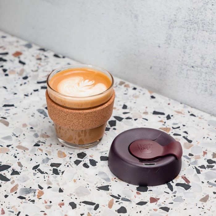 KeepCup Brew Cork XS 6oz in Alder, featuring a glass cup with cork band, filled with a flat white showing latte art, sits on a terrazzo surface beside its dark plum-coloured silicone lid.