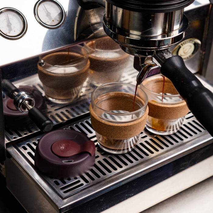 A KeepCup Brew Cork XS 6oz in Alder sits on an espresso machine drip tray as fresh espresso pours into the glass cup, featuring a natural cork band and dark burgundy lid resting beside it.