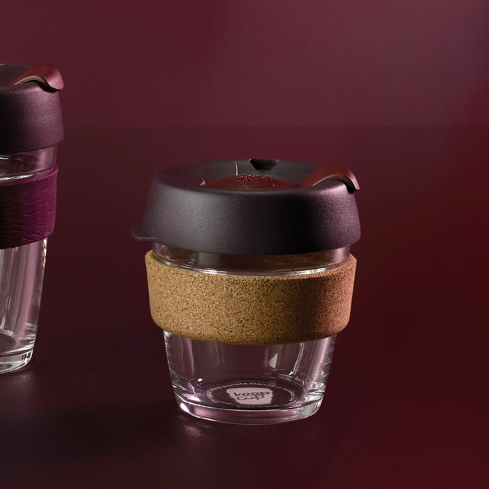 KeepCup Brew Cork XS 6oz in Alder colour, featuring a clear glass cup with natural cork band and dark charcoal lid, shown against a deep burgundy background with a second KeepCup partially visible to the left.